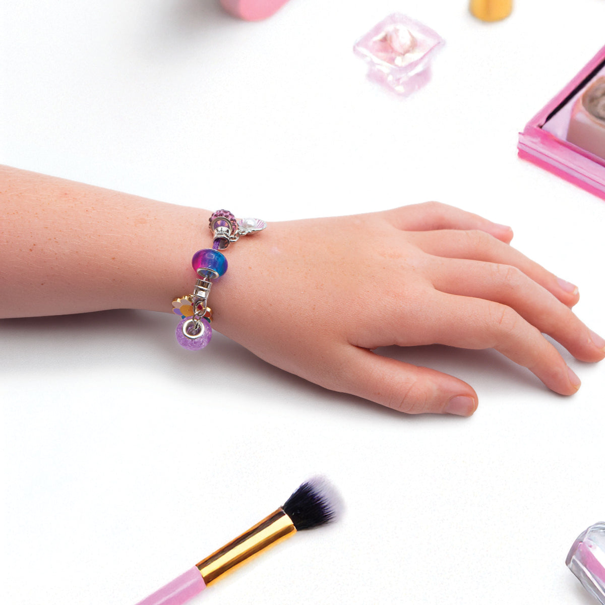 Child's hand wearing a colorful beaded bracelet with makeup brushes and a mirror in the background.